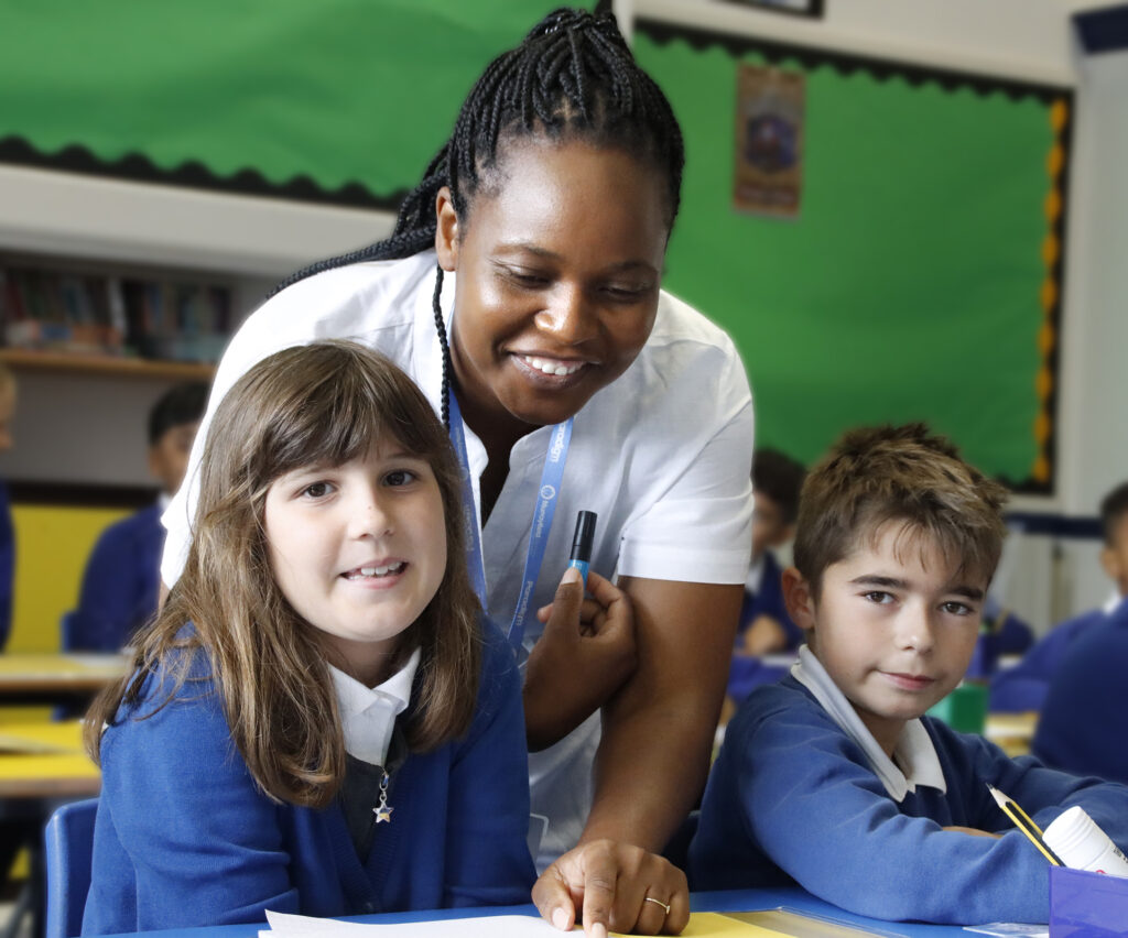 A teacher leans in between pupils to assist them with their work at the desk