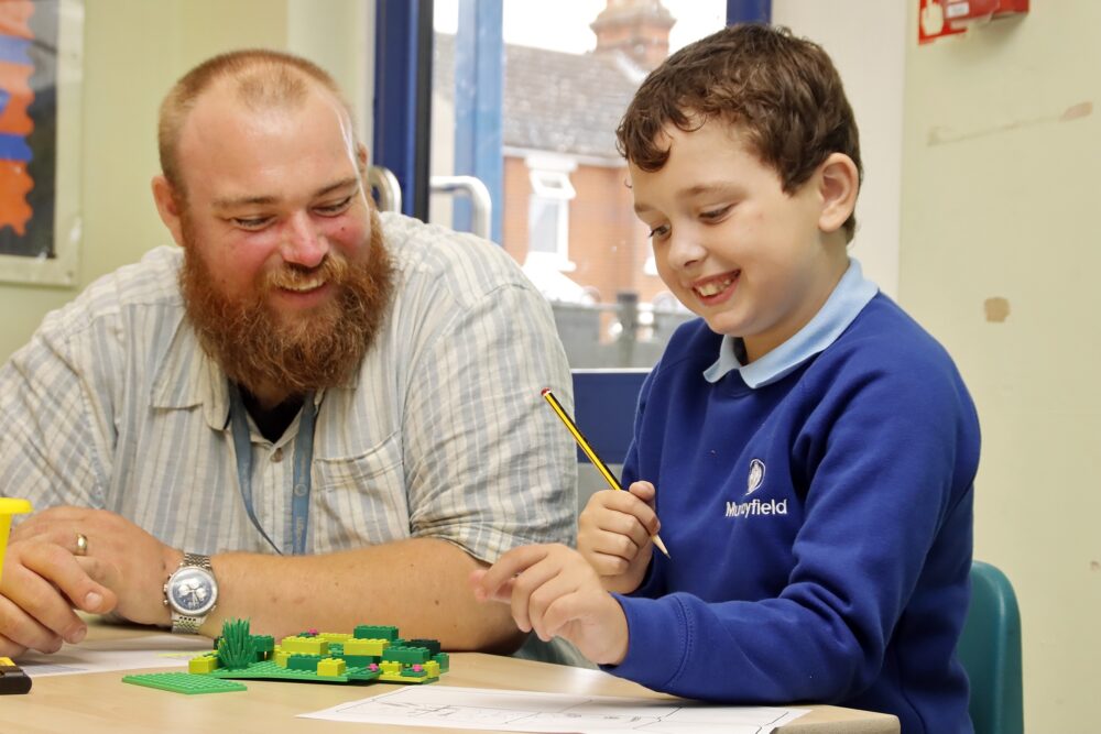 A male teacher assists a primary pupil with a puzzle using green lego bricks to solve it