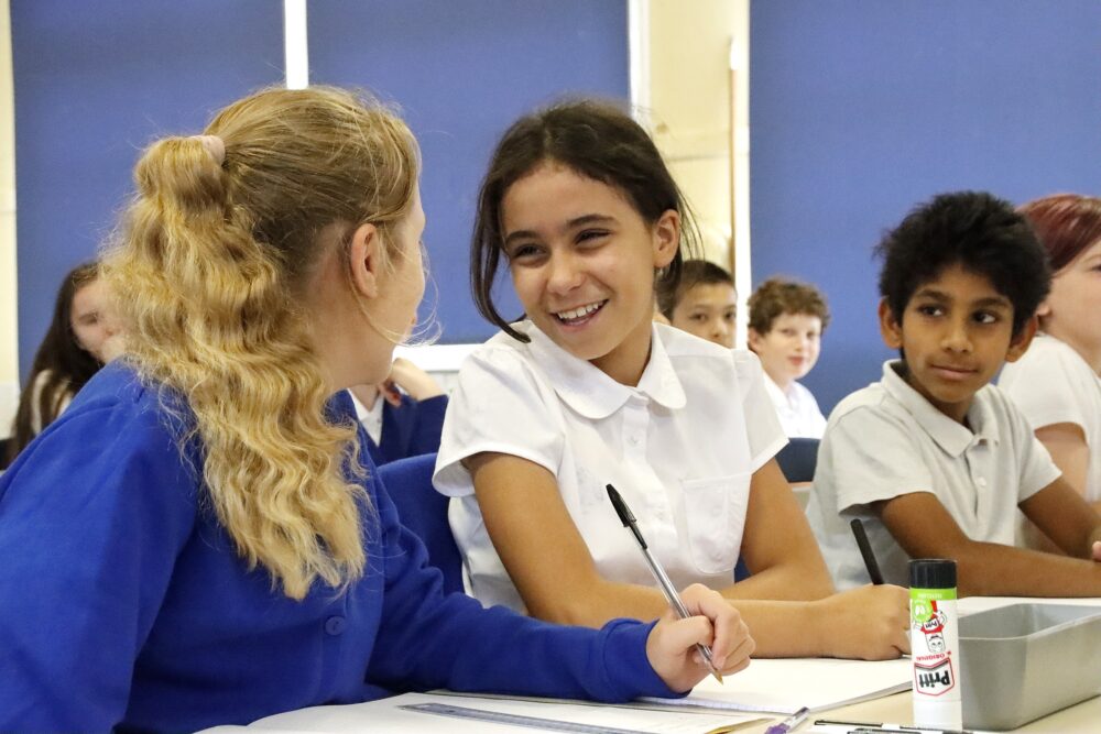 Two students working together at a desk are elated and classmates look on