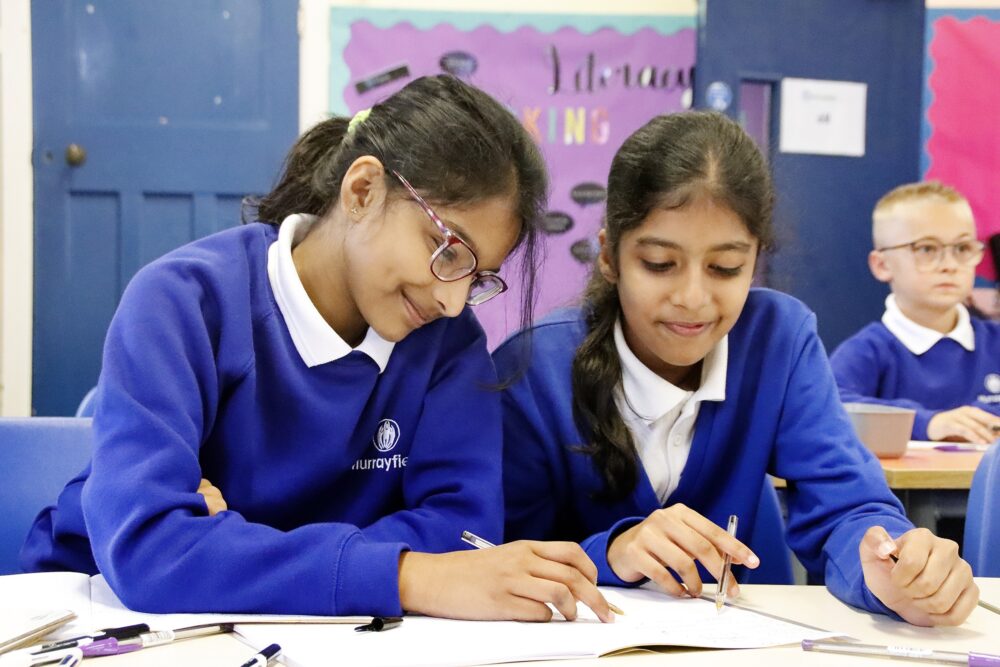 Two girl pupils work closely on a task and consider a workbook on the table in front of them