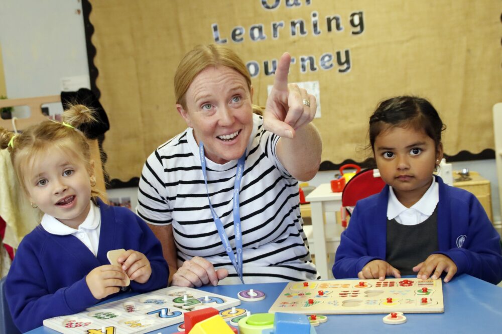 A teacher points out a feature whilst the two pupils sit either side with wooden puzzles on the table in front of them