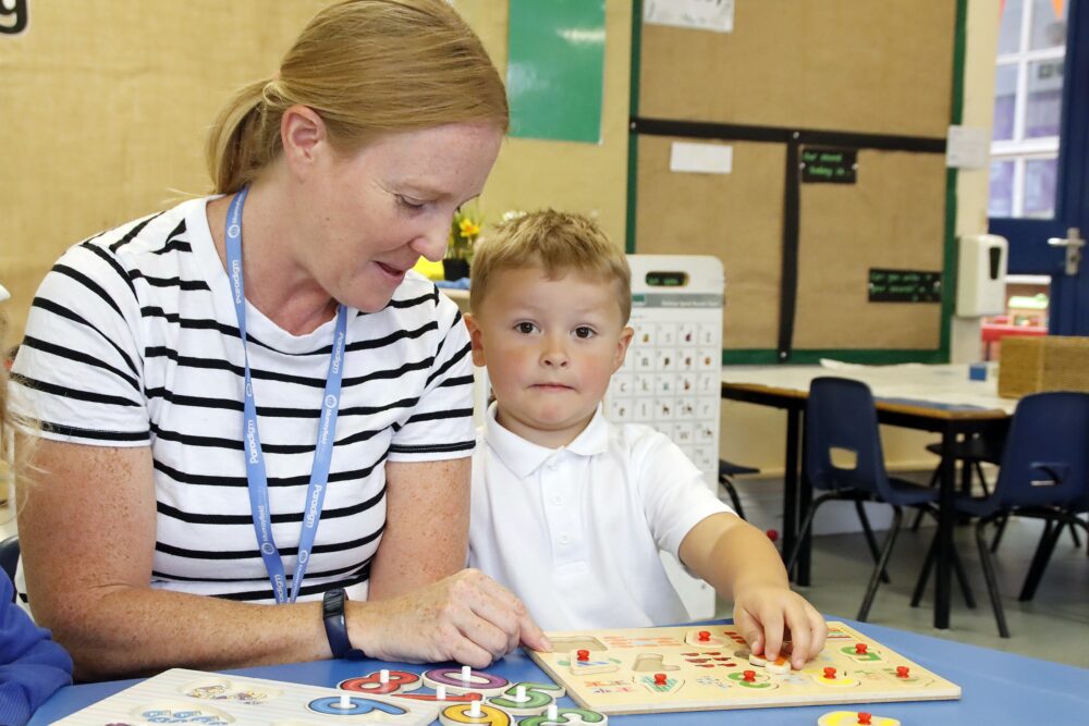 A child sits close to the teacher who assists him with a wooden number puzzle
