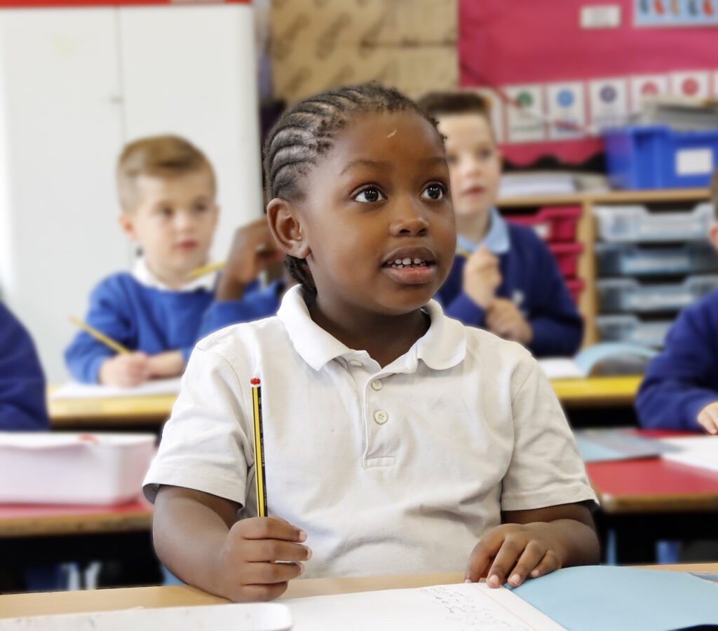A young boy eagerly looks toward the front of the classroom whilst seated at his desk, pencil in hand