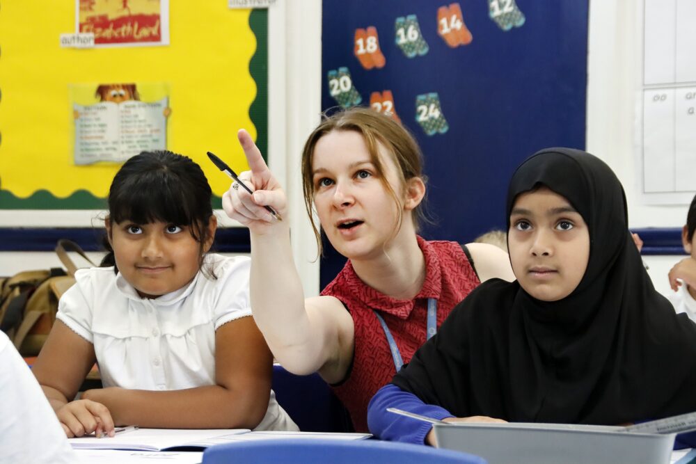A teacher crouched between two pupils focuses their attention by pointing to the board