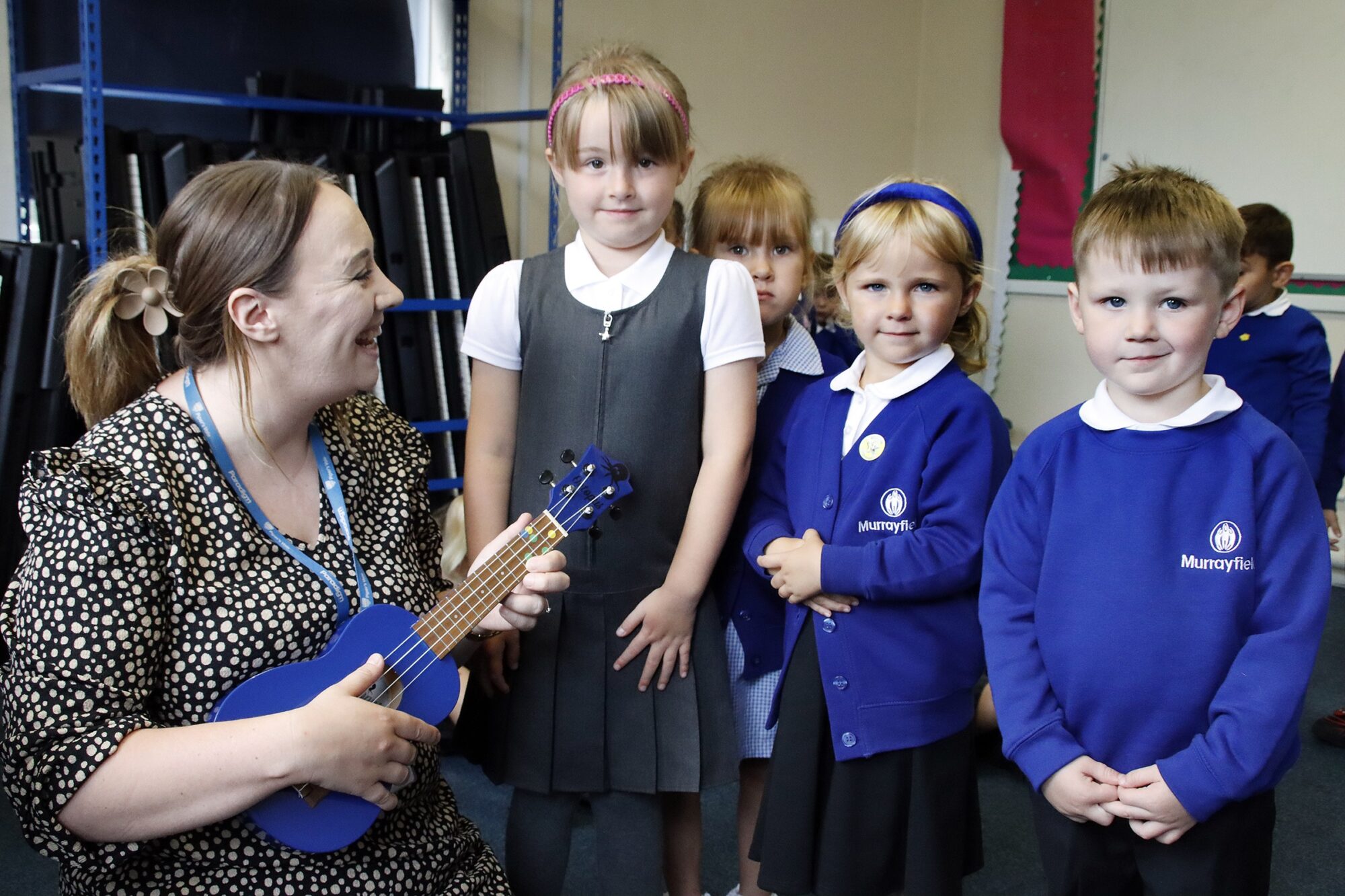 The teacher smiles whilst playing a tune on the Ukelele, the children pull a variety of faces and are full of character