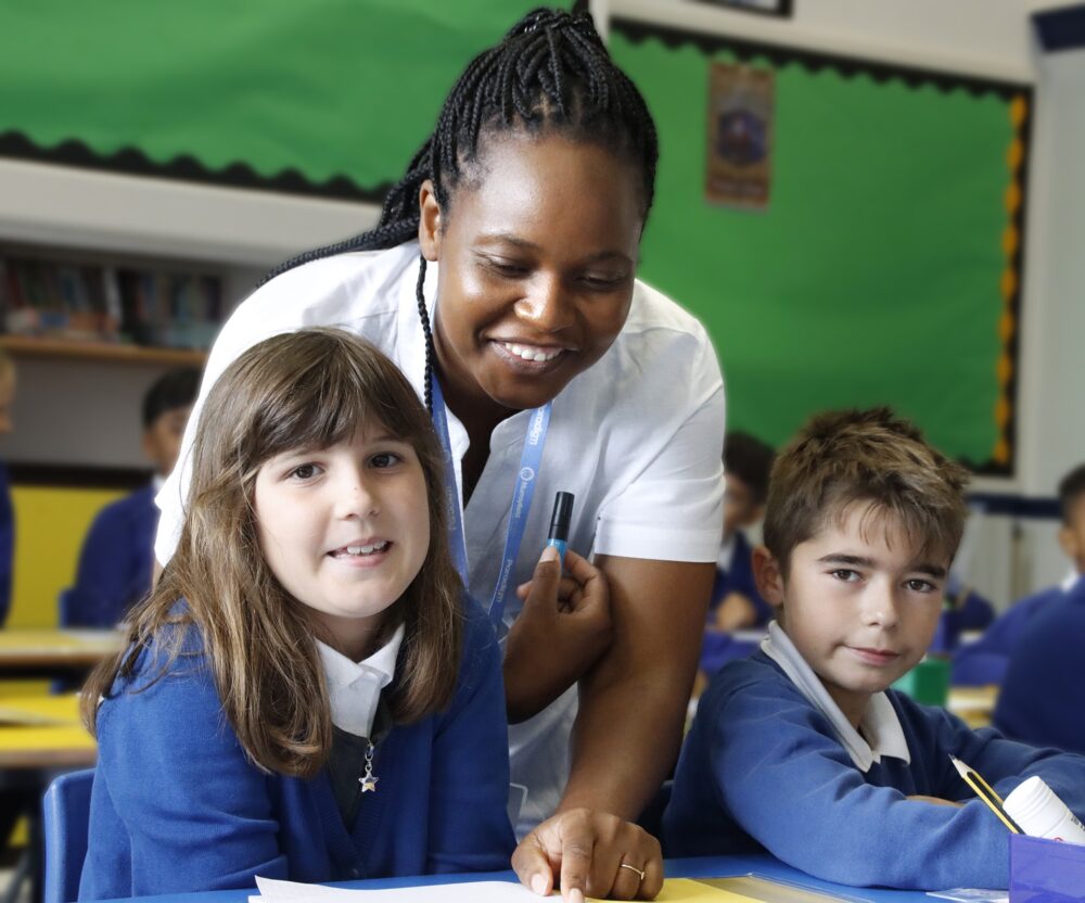 A teacher leans between two seated pupils to assist them with their project