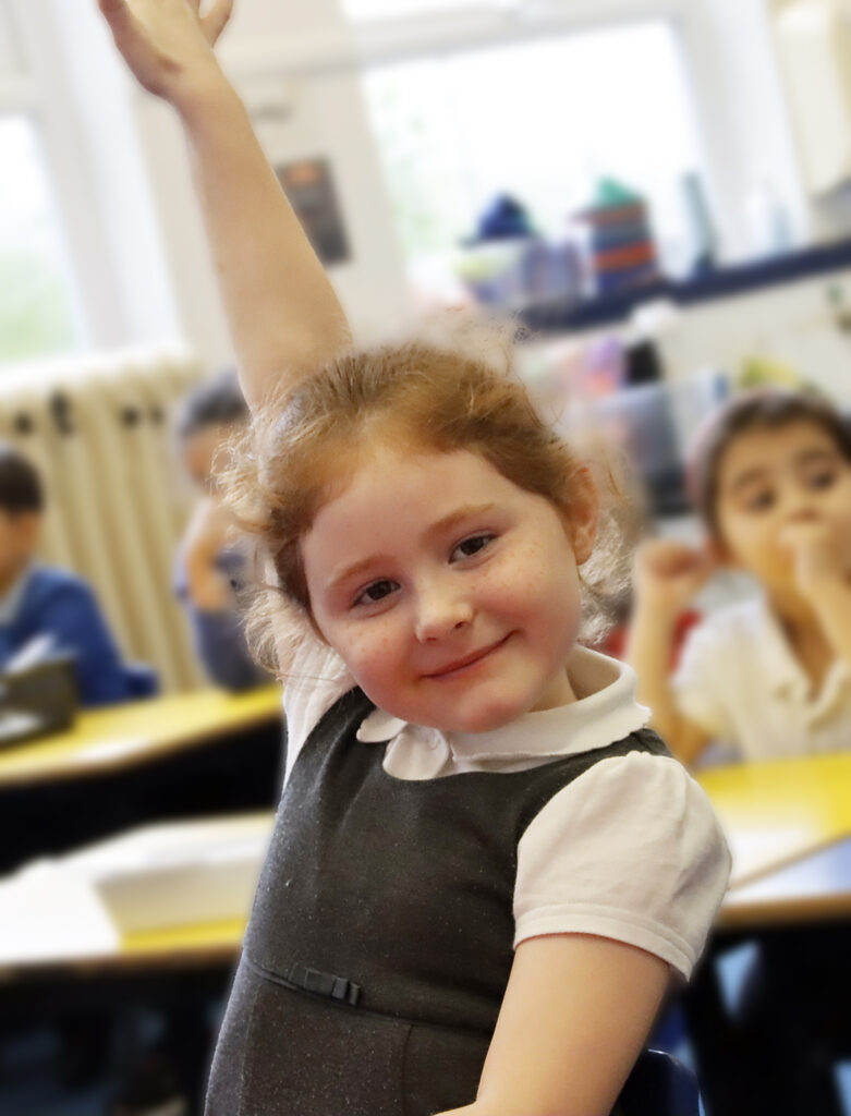 A young girl pupil holds her hand aloft and smiles ready to answer a question