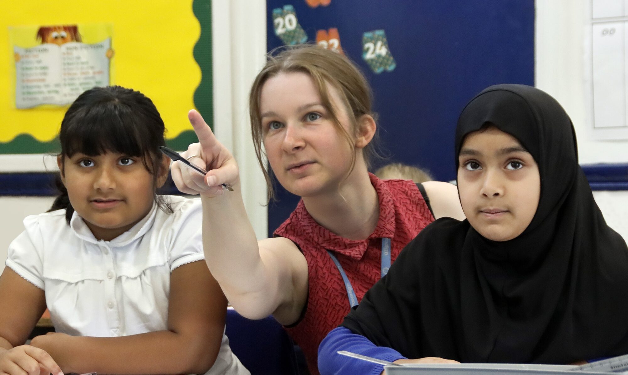 A teacher points out a mark on the wall or board for the pupils at the desk