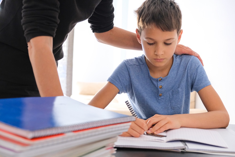 Kid sitting at the table with many books notebooks and doing homework. Mother help him. Learning difficulties, learning at home, education concept.